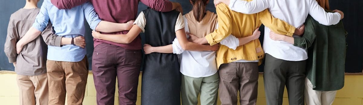 A diverse group of adults in casual outfits hugging in front of a chalkboard, symbolizing teamwork.
